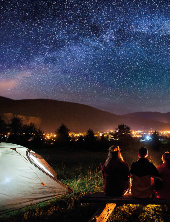 Silhouette of four people sitting on a bench made of logs and watching fire together beside camp and tents in the night  On the background starry sky, Milky way, mountains and luminous town  Rear view