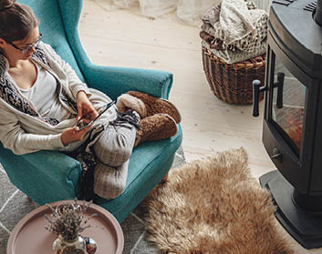 Young woman by the fireplace, sitting in a cozy armchair, with a warm blanket, using a tablet