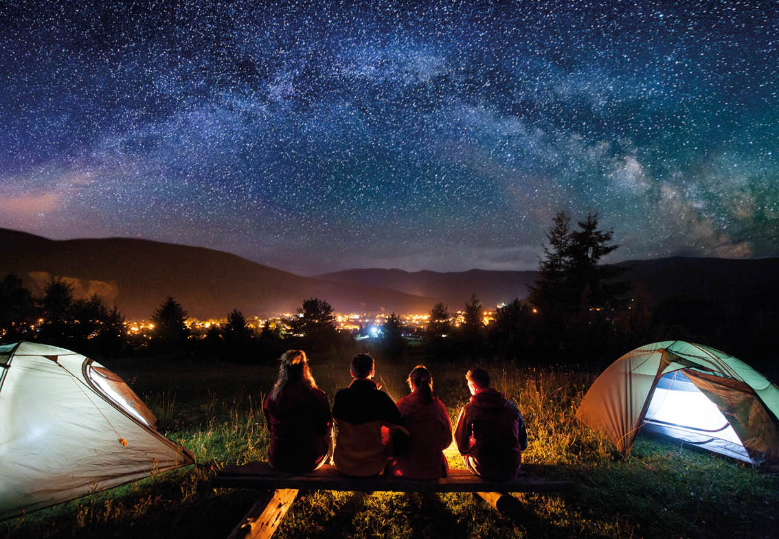 Silhouette of four people sitting on a bench made of logs and watching fire together beside camp and tents in the night  On the background starry sky, Milky way, mountains and luminous town  Rear view