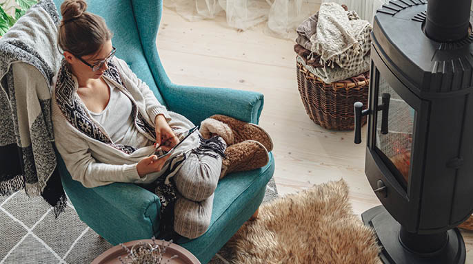 Young woman by the fireplace, sitting in a cozy armchair, with a warm blanket, using a tablet