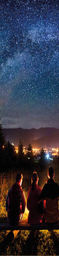 Silhouette of four people sitting on a bench made of logs and watching fire together beside camp and tents in the night  On the background starry sky, Milky way, mountains and luminous town  Rear view